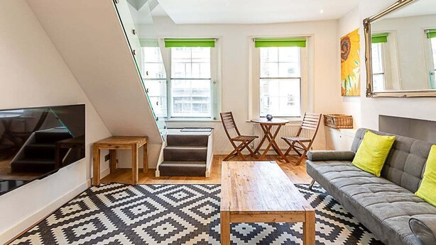 A modern loft apartment with a large window, a sofa, a dining table, and a TV mounted on the wall. - Farringdon Leo Loft (London)