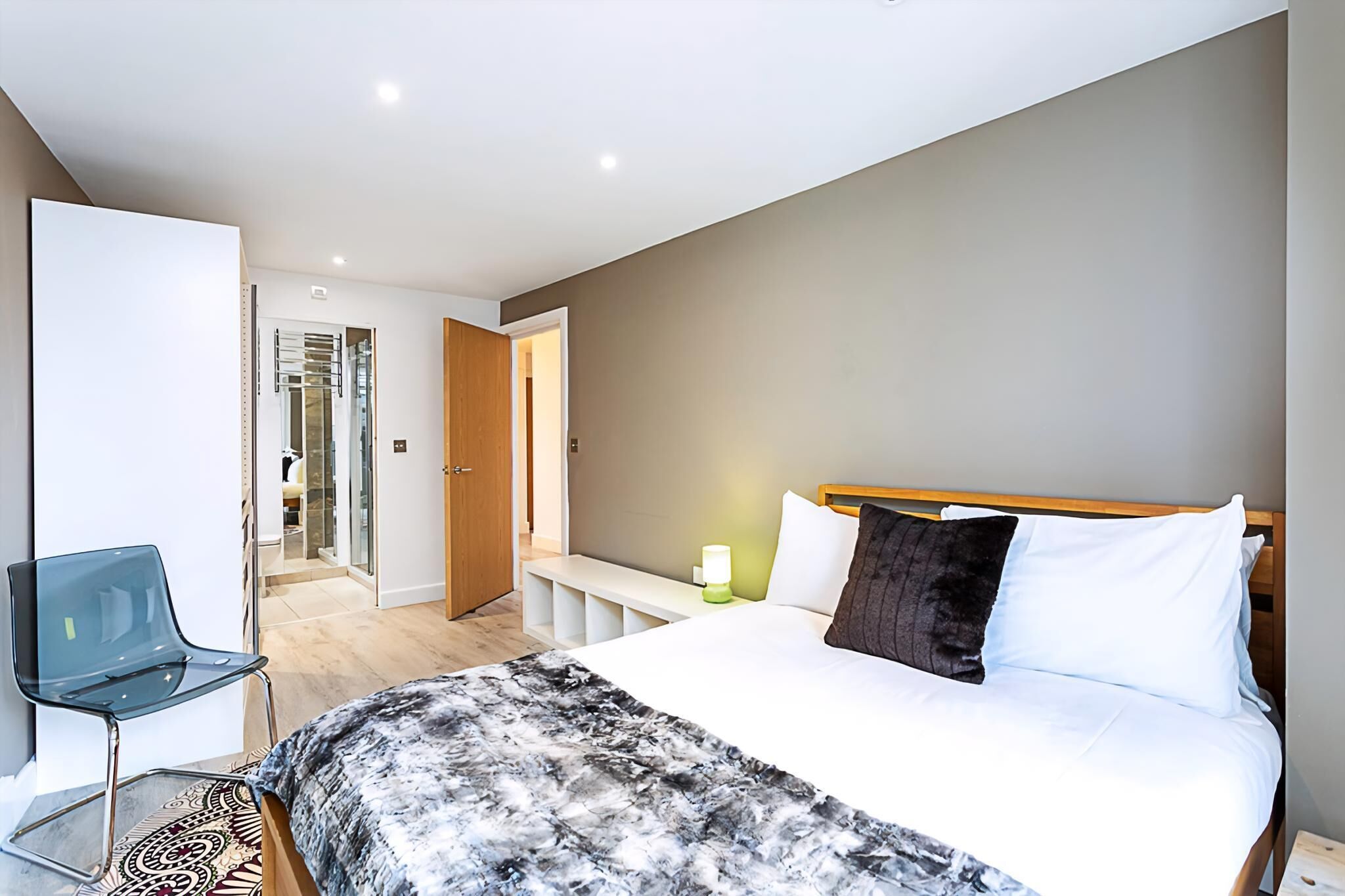 A modern hotel room with a large bed, a bedside table with a lamp, a glass chair, and a bathroom visible through an open door.