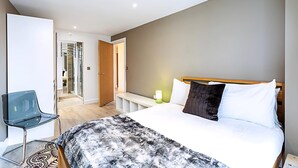 A modern hotel room with a large bed, a bedside table with a lamp, a glass chair, and a bathroom visible through an open door. - Duplex Two-bed Apartment Near Tower Bridge (London)