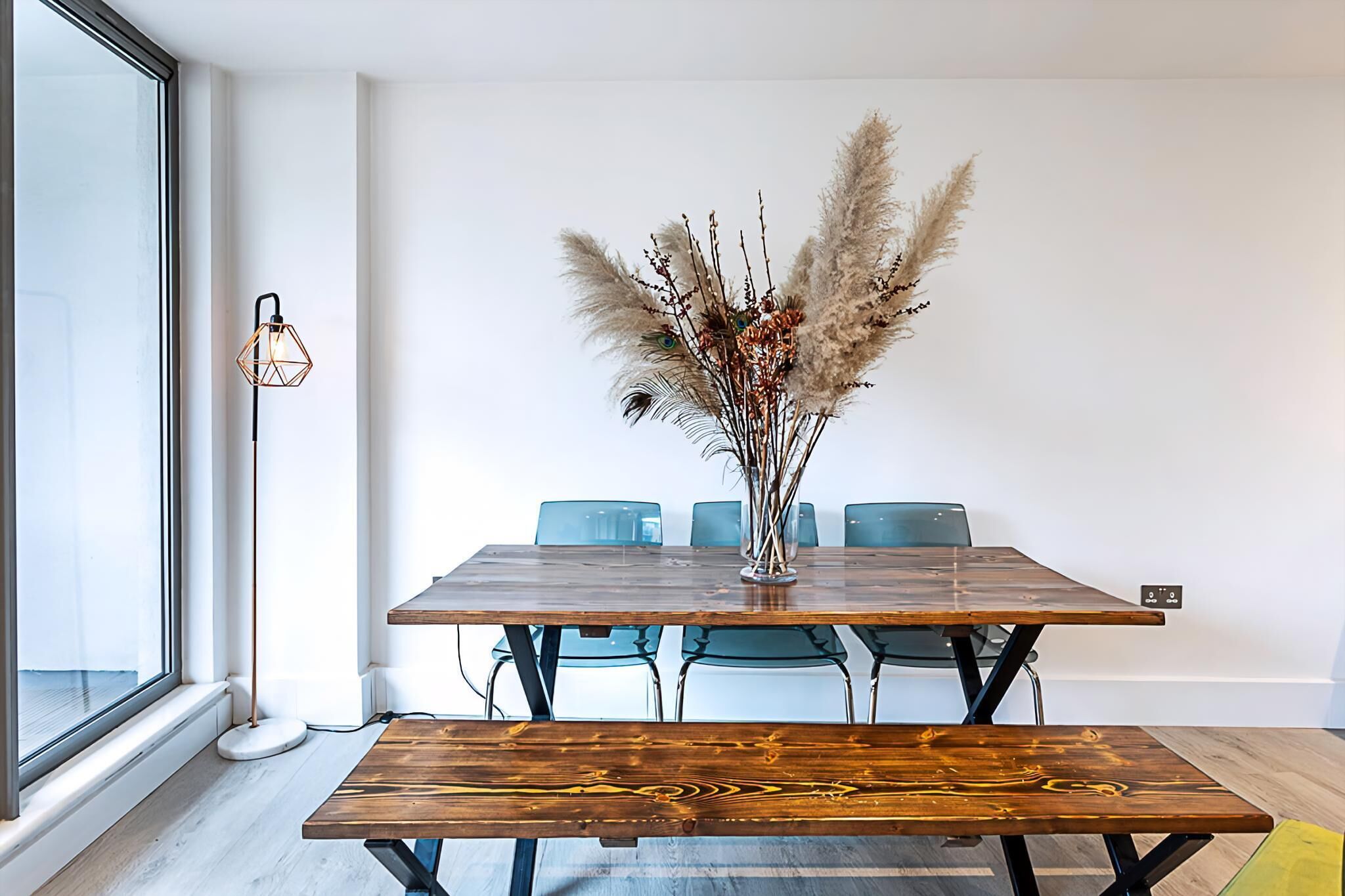 Un espace repas moderne avec une table en bois, des chaises bleues et un vase décoratif contenant des plumes d’herbe de la pampa.