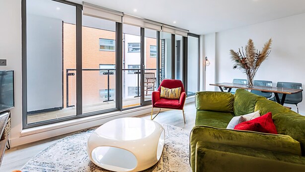 A modern living room with large windows, a red chair, a green sofa, and a white coffee table. - Duplex Two-bed Apartment Near Tower Bridge (London)