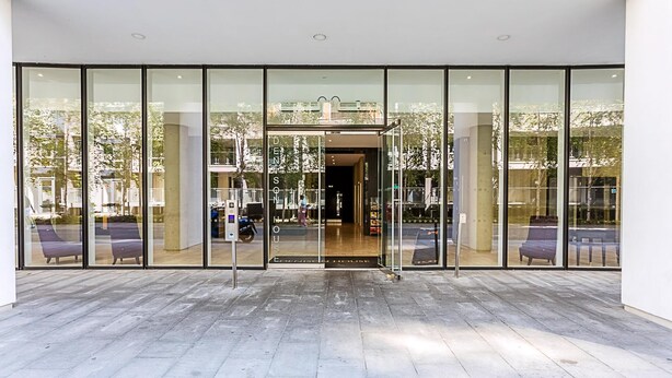 Una entrada moderna de edificio con amplias puertas de cristal, asientos exteriores y un interior visible.