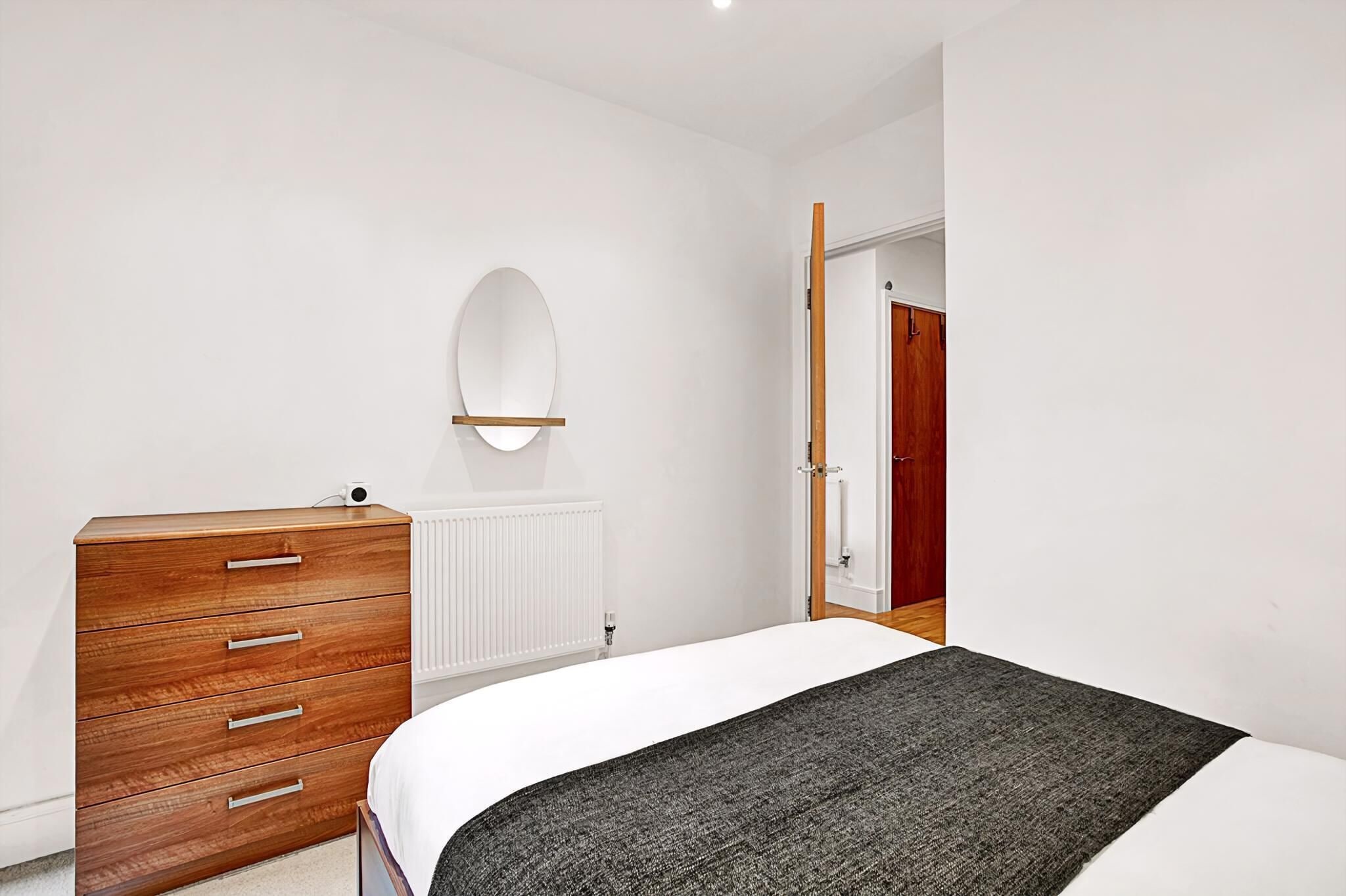 Une chambre avec une commode en bois, un lit recouvert d’une couverture grise, un radiateur blanc et une porte en bois.