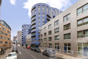 A modern urban street with multi-story residential buildings, parked cars, and a clear sky.