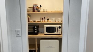 A compact kitchen with a microwave, toaster, and refrigerator, featuring a wooden shelf with kitchenware and a small basket.