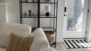 A living room with a white sofa, a black metal shelf with decorative items, a white door, and a view of the outside.