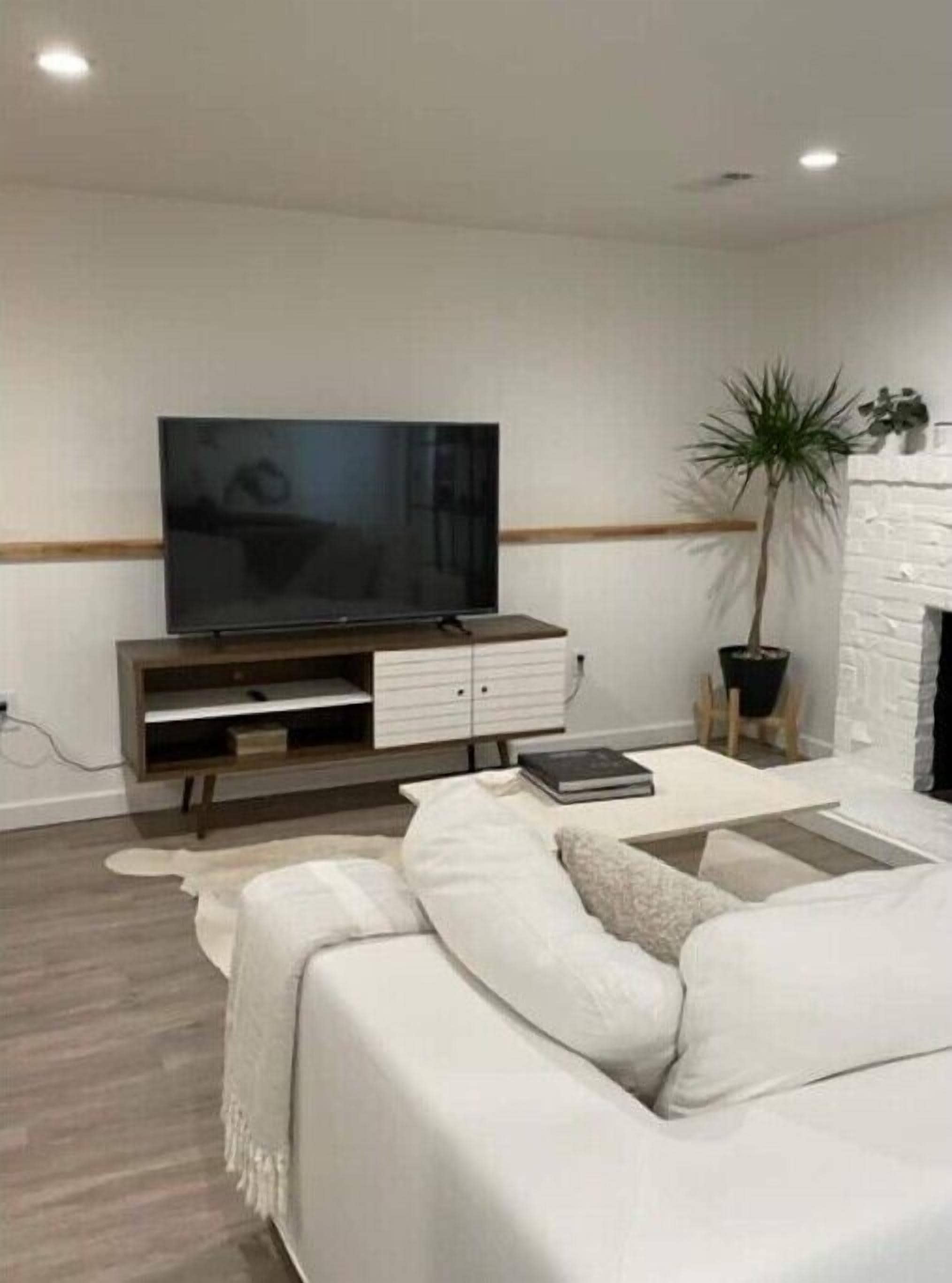 Un salon moderne avec un téléviseur à écran plat posé sur un meuble en bois, un canapé blanc, une petite table d’appoint et une plante en pot.