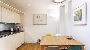A modern kitchen with white cabinets, a black countertop, and a wooden dining table with chairs. There are framed artworks on the walls and a floor lamp. - Perfect Paddington Pad (London)
