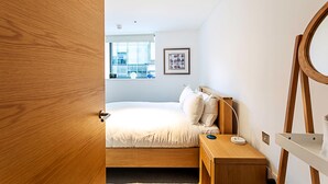 A bedroom with a bed, bedside table, lamp, and a window with a view of the outside. - Perfect Paddington Pad (London)