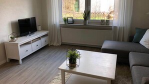 Ein modernes Wohnzimmer mit einem Flachbildfernseher auf einem weißen TV-Schrank, einem grauen Sofa, einem weißen Couchtisch und einem Fenster mit Blick auf einen Garten.