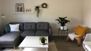Ein Wohnzimmer mit grauem Ecksofa, einem weißen Couchtisch, einem weißen, flauschigen Teppich, einem kleinen Beistelltisch mit Pflanze und einem Korbstuhl mit gelbem Kissen.