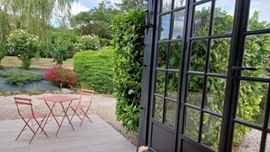 Pátio com mesa e cadeiras, cercado por vegetação exuberante e vista através de uma grande janela.