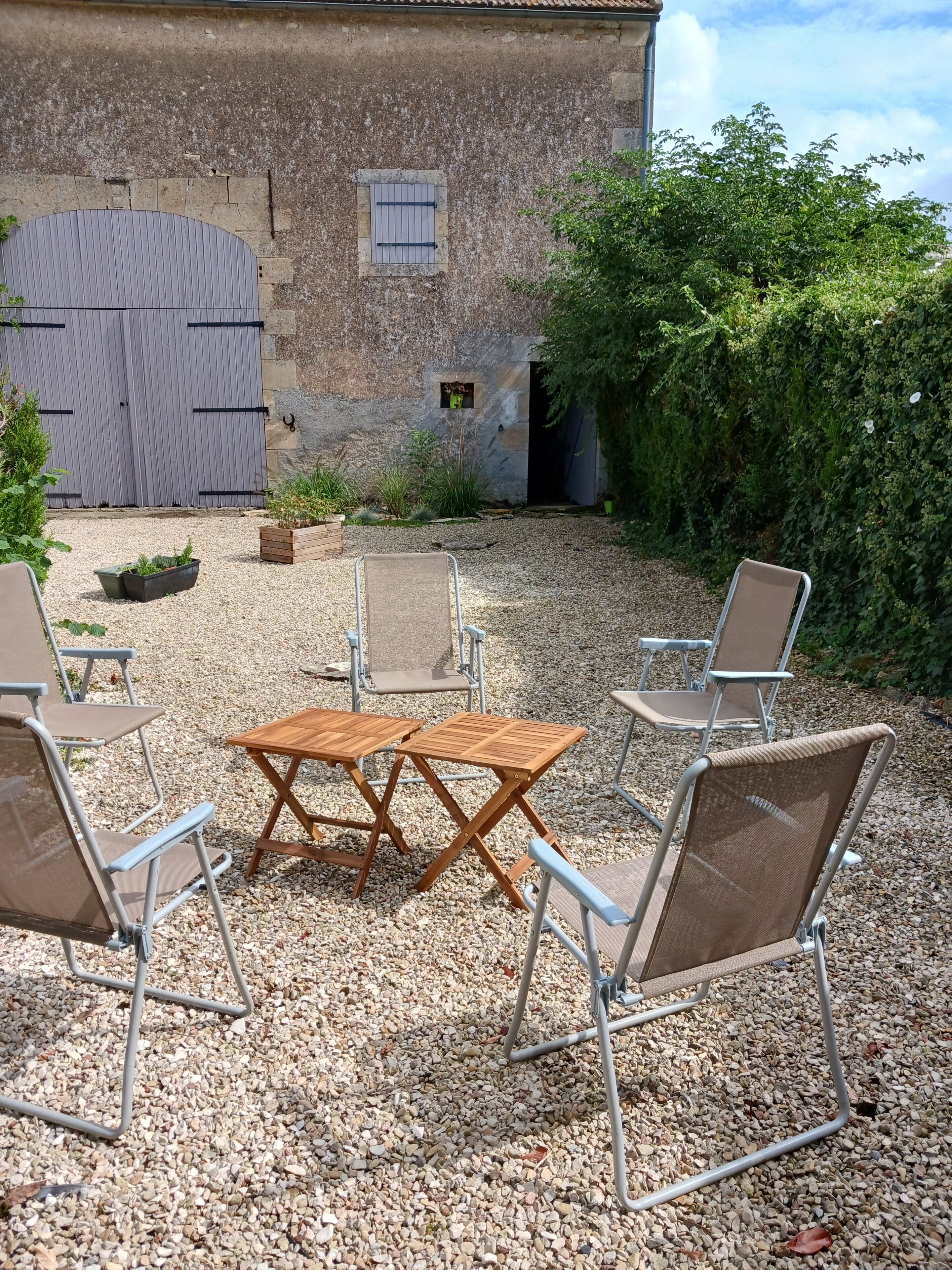An outdoor seating area with a wooden table and metal chairs, gravel ground, and a stone building with a closed shutter in the background.