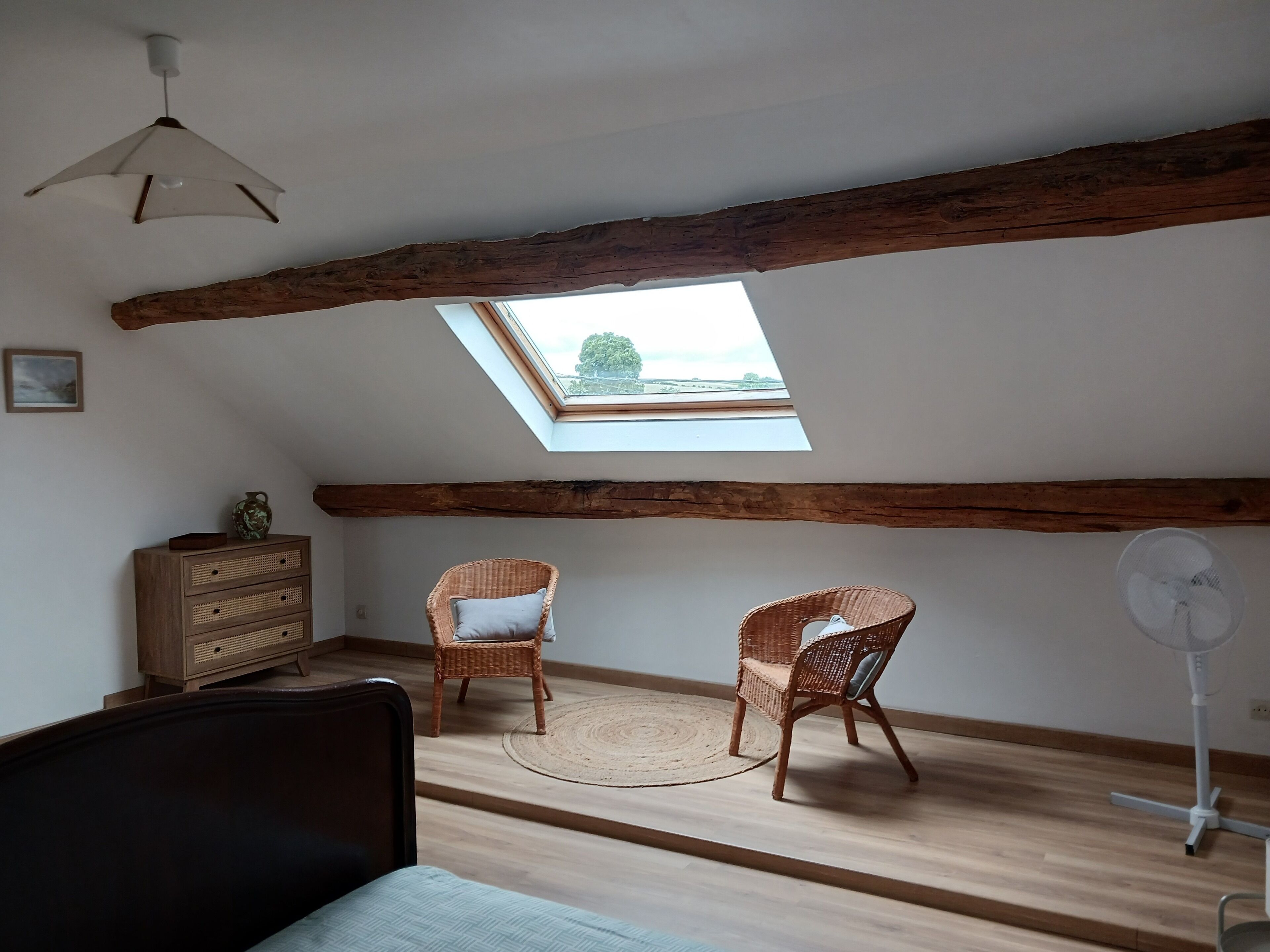 Une pièce avec un plafond en pente, des poutres en bois, deux fauteuils en osier, une petite armoire en bois et un ventilateur sur pied.