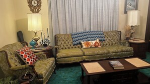 A living room with a green sofa, a patterned armchair, a coffee table, and a side table with a lamp and decorative items. - Groovy 1970s Home in central Indiana with basement full of entertainment! (Carmel)