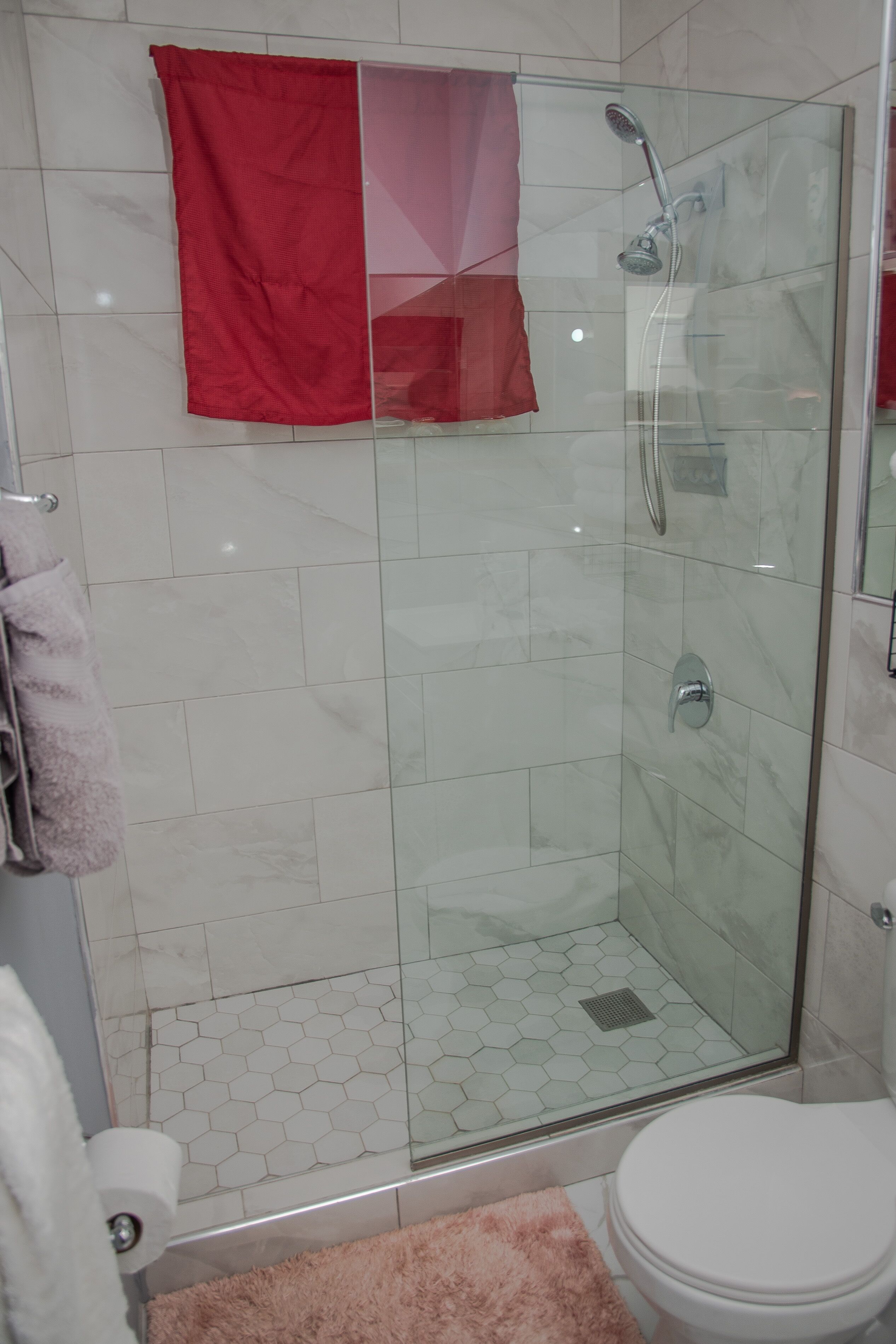 A glass-enclosed shower with a handheld showerhead, a toilet, and a pink rug.