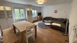 A modern living room with a sofa, a dining table, and a kitchen area with cabinets and a radiator. - Ruhig und Idyllisch: Kleines Ferienhaus am Waldrand - S-bahn-nähe - Zentrumsnah (Bergisch Gladbach)