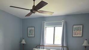 A bedroom with a bed, a ceiling fan, a window with curtains, two bedside tables with lamps, and two framed pictures on the wall.