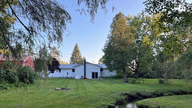 A white building with a chimney, surrounded by a grassy yard and a small stream.