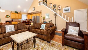 A spacious living area with a brown leather sofa, a matching armchair, and a coffee table. A staircase with a white railing leads to an upper level.