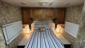 A compact RV bedroom with a striped bed, wooden cabinets, and two wall-mounted lights. - Cozy little camper, nestled quietly in the country / only minutes from the city  (Purvis)