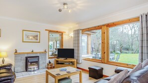 A living room with a fireplace, a television, a sofa, a coffee table, and large windows with a view of trees. - 5 bedroom accommodation in Nethy Bridge (Nethy Bridge)