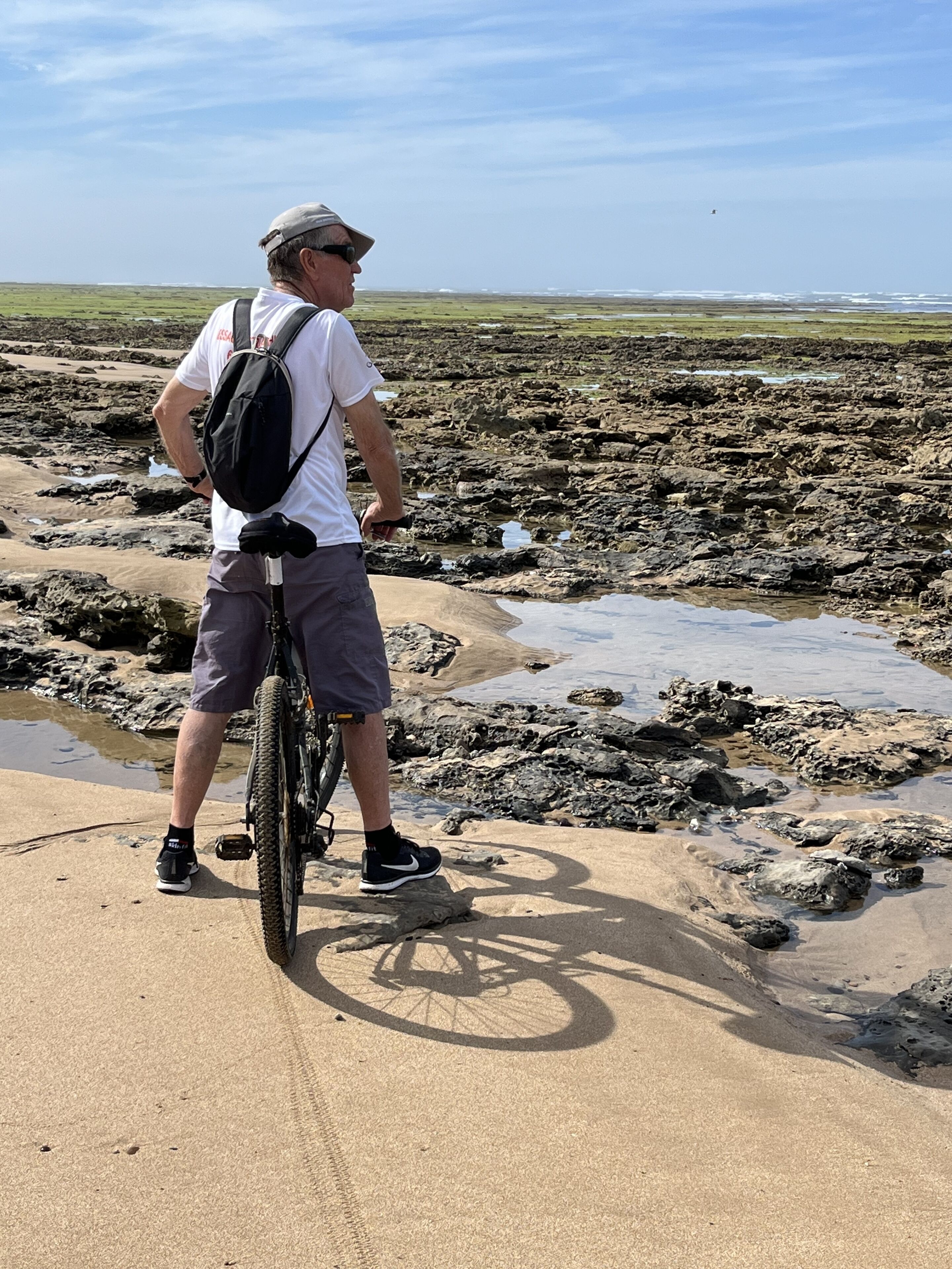 Una persona in piedi accanto a una bicicletta su una spiaggia rocciosa, con un cielo sereno sullo sfondo.