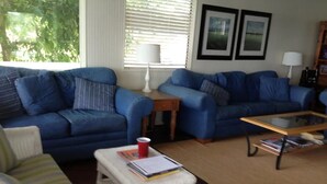 Una sala de estar con dos sofás azules, un sillón blanco, una mesita pequeña con una taza roja y una mesa de centro con superficie de vidrio y libros.