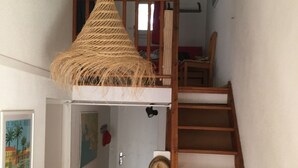 Een smalle gang met een houten ladder, een hanglamp van geweven materiaal en een deur die leidt naar een slaapkamer.
