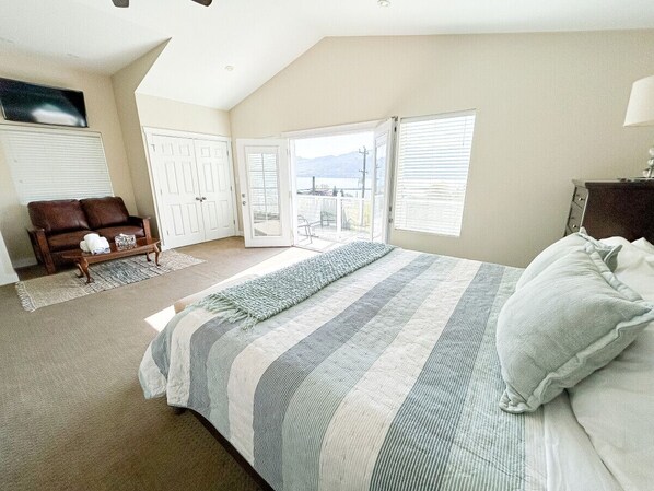 A bedroom with a large bed, a view through the open door, a wooden bench, and a television on the wall.