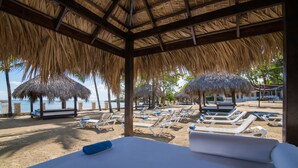 Un cabanon de plage avec un toit de chaume, des chaises longues blanches et un lit avec des serviettes bleues.