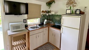 Een kleine keuken met een magnetron, spoelbak en koelkast. Er zijn houten balken, een flatscreen televisie en een klein raam.