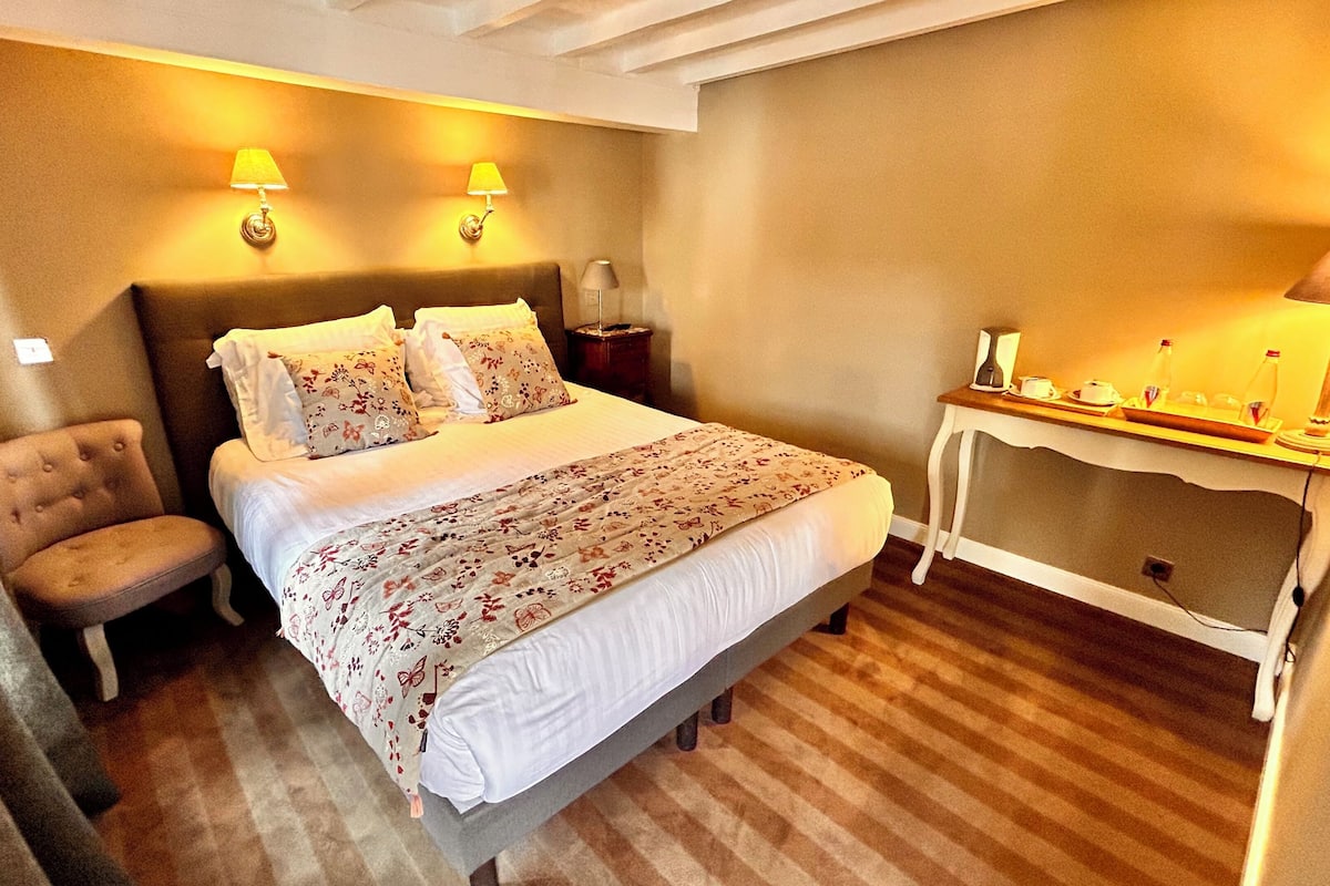 A hotel room with a bed, a chair, a nightstand, a table with a lamp, and a small table with a lamp and a tray of items.