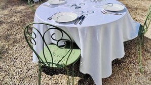 Un área de comedor al aire libre con mesas redondas cubiertas con manteles blancos, sillas de metal y un muro de piedra de fondo.