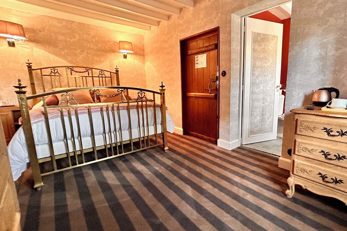 A hotel room with a bed, a wooden dresser, a door, and a wall-mounted lamp.