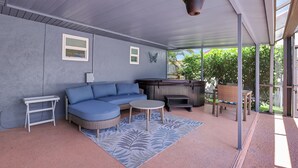 A covered patio area with a blue sectional sofa, a small round table, a white side table, a hot tub, and a hanging plant. - Pure Living, Saltwater Pool, Low Toxin Getaway! (Port St. Lucie)