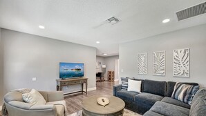 A spacious living room with a grey sofa, a round coffee table, and a beige armchair. There is a painting on the wall and a desk with a lamp. - Pure Living, Saltwater Pool, Low Toxin Getaway! (Port St. Lucie)