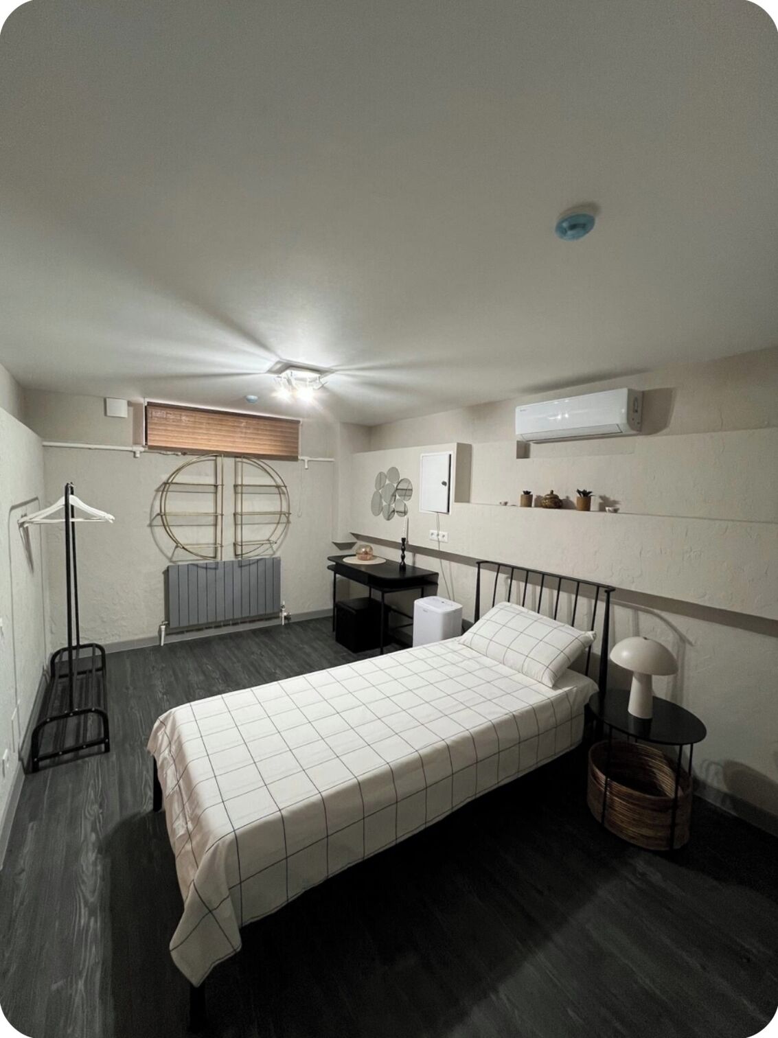 Een kleine slaapkamer met een bed, nachtkastje, lamp, radiator en een klein bureau met stoel.