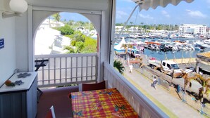 Uma varanda com toalha de mesa xadrez, uma mesa pequena e uma cadeira. Uma vista da marina, com barcos e edifícios, é visível pela janela.