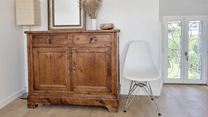 Un aparador de madera con un espejo y flores secas, una silla blanca y una puerta con vistas a la vegetación.