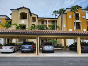 A parking structure with multiple cars parked under a covered area, adjacent to a yellow building with balconies and palm trees.