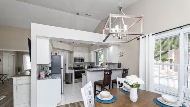 A spacious kitchen with white cabinetry, a central island, and a dining area with a wooden table set for four. - Castle Glow - 2BR - BBQ Gr - Walk to Kroger Field (Lexington)