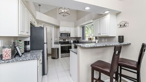 A modern kitchen with white cabinetry, a granite countertop, stainless steel appliances, and a bar area with two wooden chairs. - Castle Glow - 2BR - BBQ Gr - Walk to Kroger Field (Lexington)