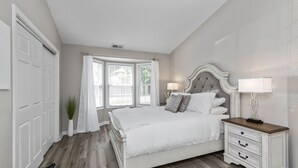 A bedroom with a white bed, a nightstand, a window with curtains, and a potted plant. - Castle Glow - 2BR - BBQ Gr - Walk to Kroger Field (Lexington)