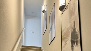 Un escalier avec des marches en bois et des rampes blanches, menant à une pièce aux murs blancs et ornée de tableaux encadrés.