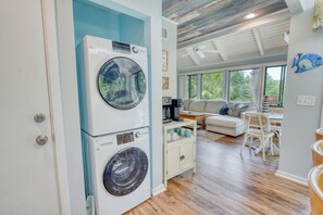Un cuarto de lavandería con lavadoras apiladas, un comedor con mesa y sillas, y una sala de estar con sofá y ventanas amplias.