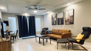 A living room with a brown leather sofa, a black armchair, a coffee table, and a ceiling fan. There are three framed pictures on the wall, a window with blinds, and a door with a curtain. - Best 3-Story Home in Vegas - 9mins to Everything! (Las Vegas)