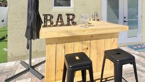 Een houten toog met de letters 'BAR', twee zwarte barkrukken en een klein tafeltje met een glas erop.
