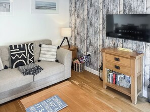 Uma sala de estar com um sofá cinzento, almofadas estampadas, uma mesa lateral de madeira com uma luminária, uma TV e uma estante de livros.
