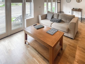 Ein Wohnzimmer mit einem Holztisch, einem beigefarbenen Sofa mit gemusterten Kissen und Blick durch große Fenster.